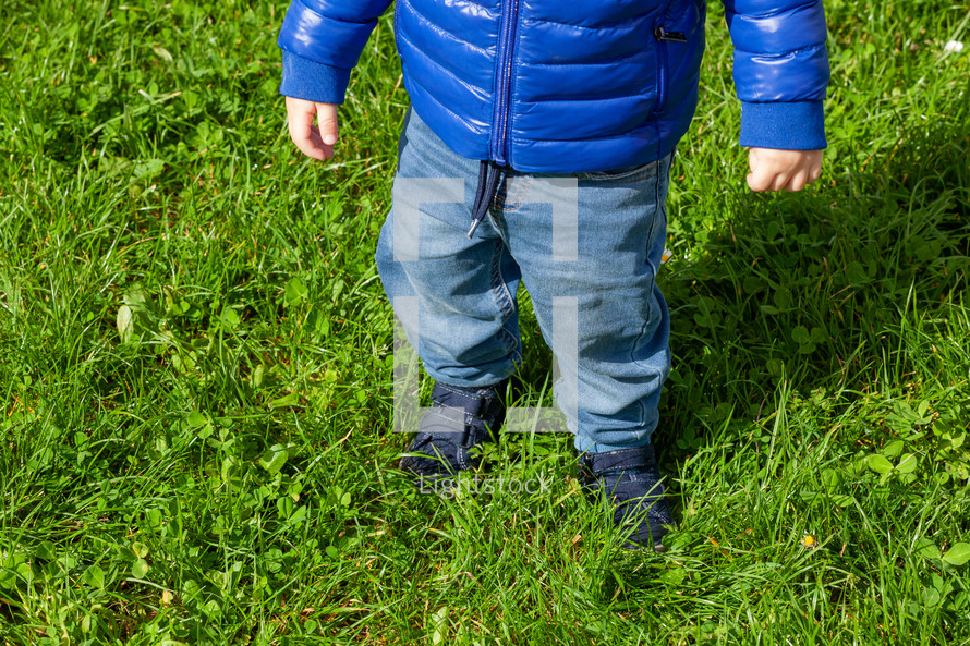 Close-up of a young child in blue taking tentative steps outdoors on vibrant green grass