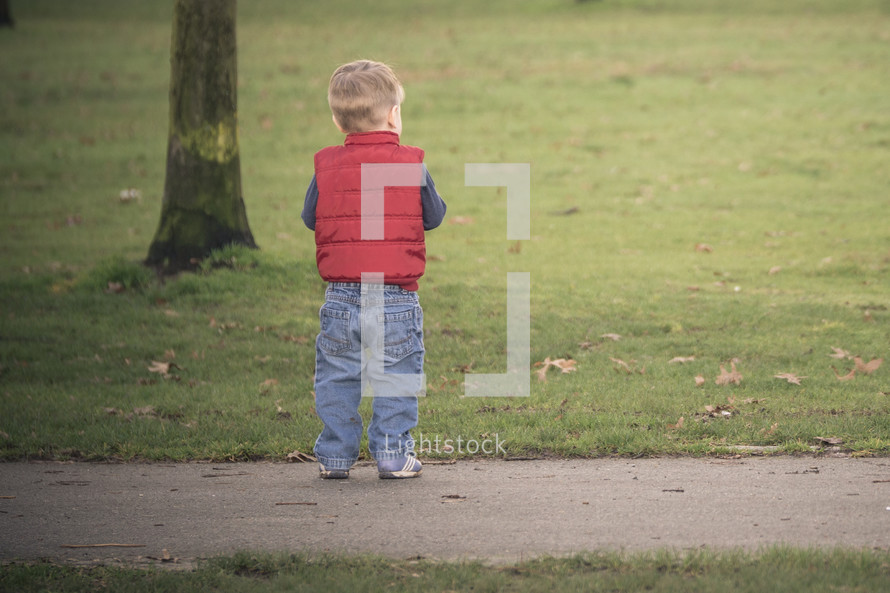toddler boy in a vest standing on a sidewalk 