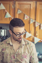 a man in a floral pattern shirt and reading glasses standing in a kitchen 