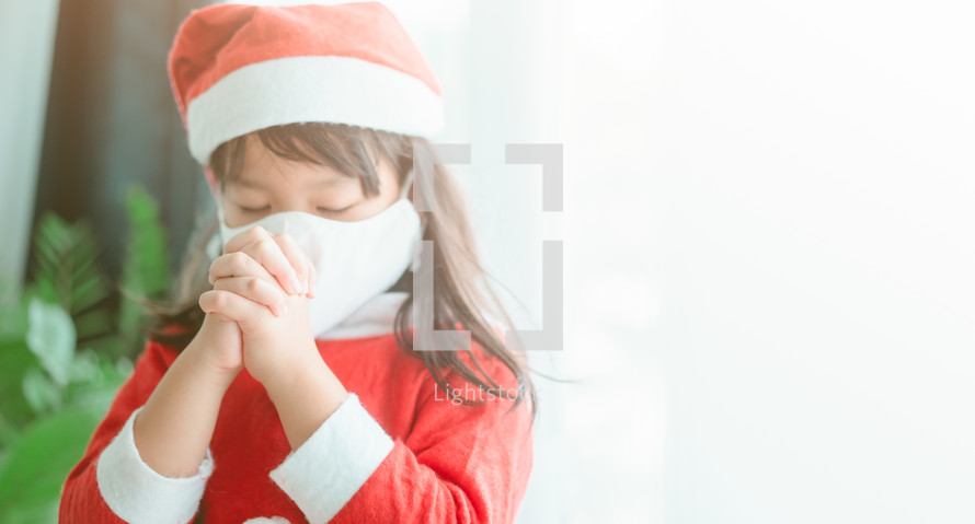 a little girl in a Santa suit praying while wearing a face mask 