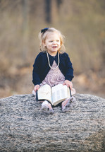 a little girl sitting on a rock holding a Bible 