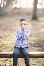 a little boy sitting on a bench outdoors praying 
