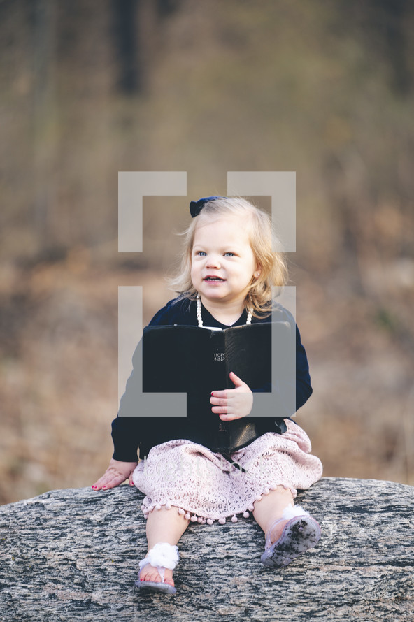 a little girl sitting on a rock holding a Bible 