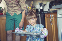 toddler girl with oven mitts carrying a muffin pan
