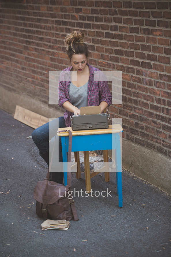 A young woman sitting at a school desk with a typewriter by a brick wall.