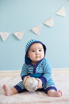 Infant boy wearing a hoodie, holding a ball, sitting on the floor.