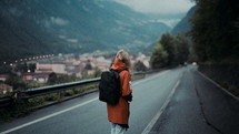 a young woman in a trench coat and backpack standing in the middle of a road 