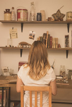 A woman sits at a craft desk with shelves on the wall.