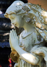 A side profile view of a statue of a praying female angel figure with a smiling face and prayerful pose adorns a courtyard in a church cemetery. 