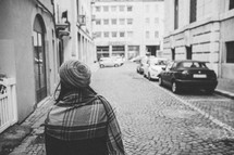a woman in a coat and shawl walking along a narrow street window shopping 