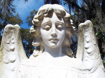 A frontal pose of an angel face and wings carved out of stone with a blue sky and trees in the background. 