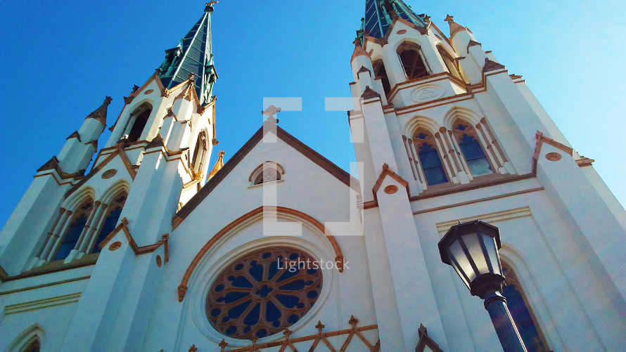 An old historic catholic church in Savannah, Georgia with large stained glass windows and gas lights stretch out into the blue sky with high arches and old gothic style architecture in the heart of the city where old and young alike can come and worship God together. 