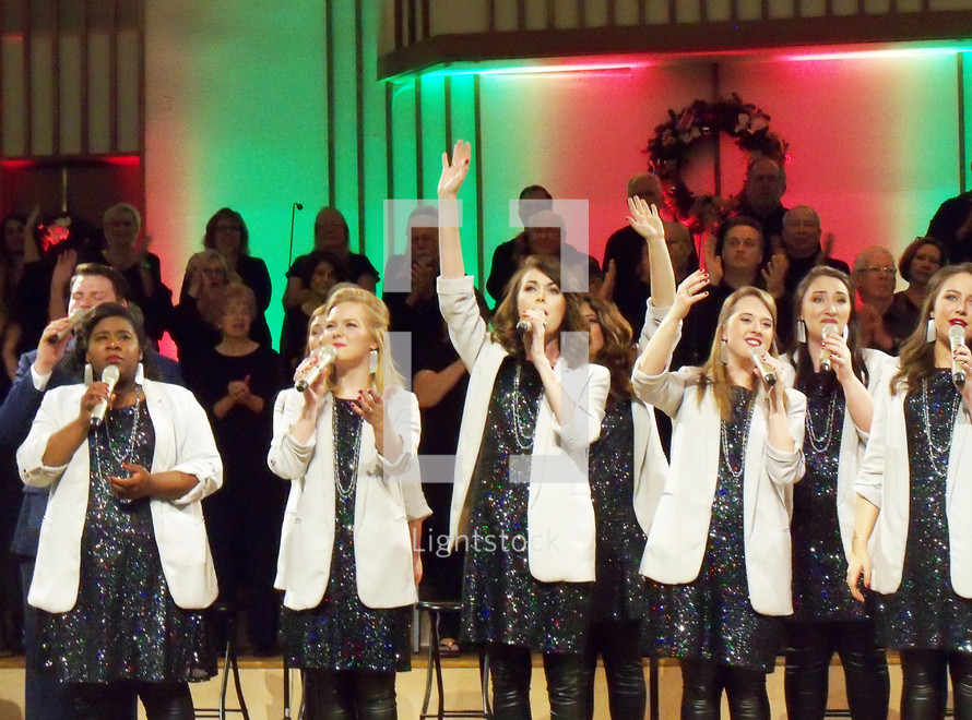 A group of female and male singers sing a chorus of praise during a Christmas praise and worship concert wearing white blazers and sparkling dresses while singing during a church Christmas concert. These are the voices of Mobile - a praise and worship group from Mobile Alabama singing at a Christmas concert along with the church choir at a recent praise and worship event. 