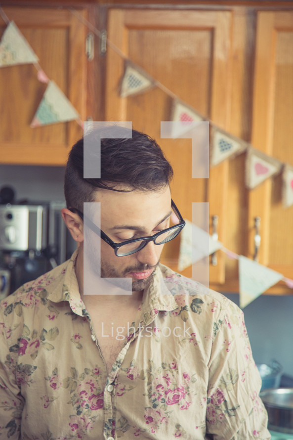a man in a floral pattern shirt and reading glasses standing in a kitchen 