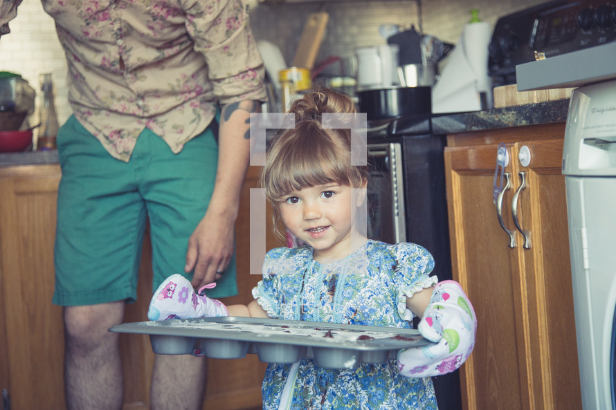 toddler girl with oven mitts carrying a muffin pan