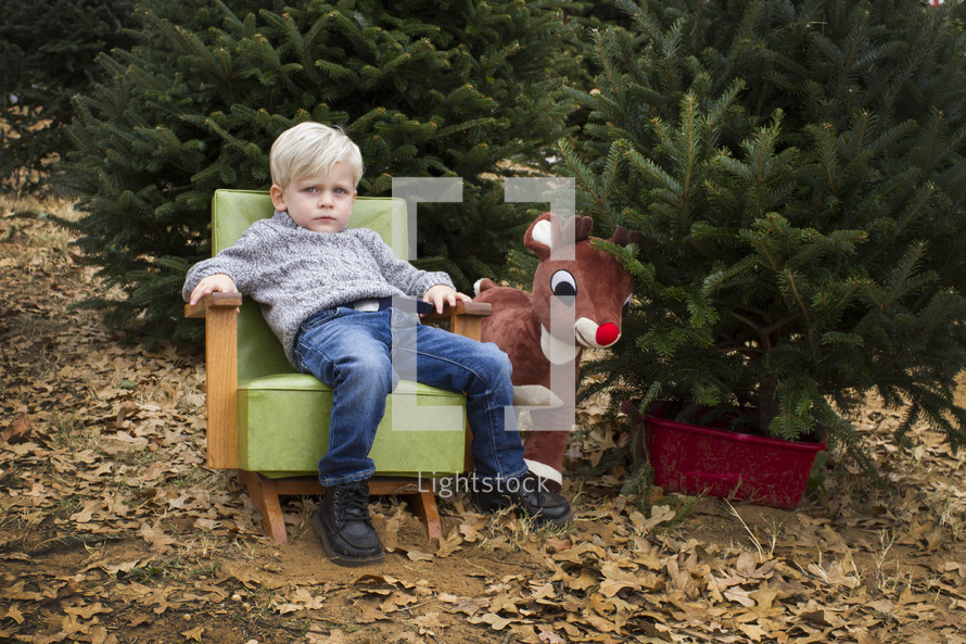 a boy child sitting in a chair next to Rudolph in a Christmas tree lot 