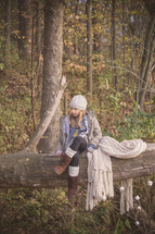 a girl with a blanket sitting on a fallen tree in a forest 