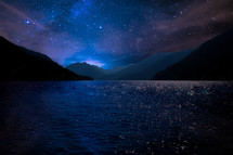 stars in the nights sky and water in a bay surrounded by mountains 