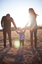 A white mom and dad help their child walk in a field.