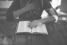 A woman reading the Bible at a table with her hand over her heart