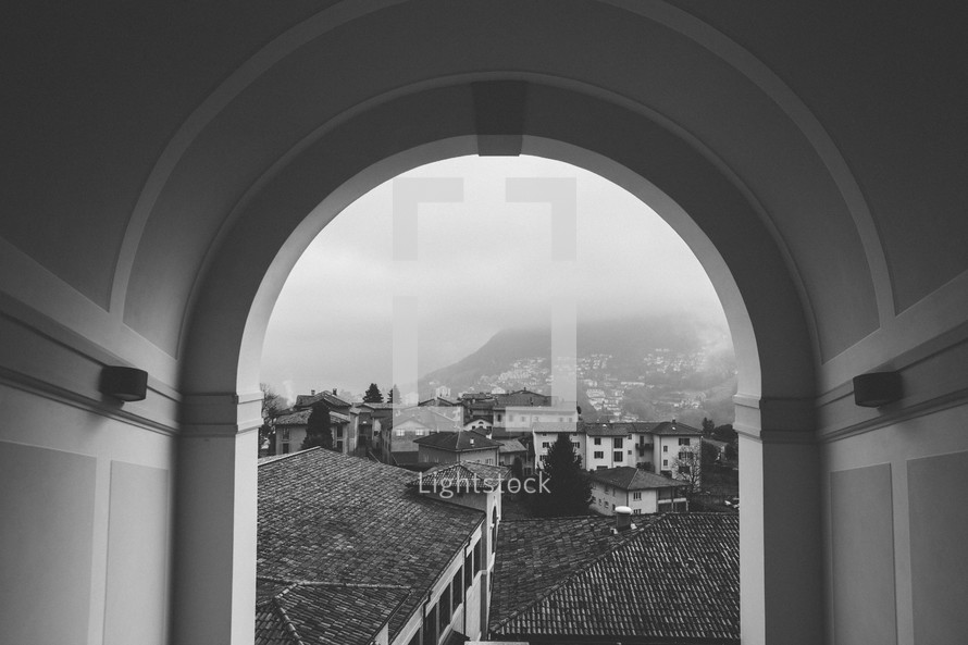 View through an arch of tile roofs, stucco buildings and  foggy mountains.