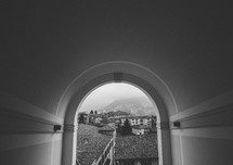 View through an archway of tile roofs, stucco buildings and foggy mountains.