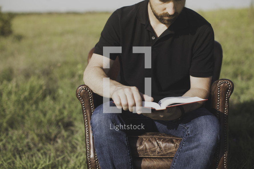a man sitting in a leather chair reading a Bible in a field 