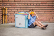 a child with a camera sitting by a locker 