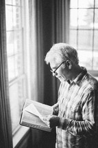 elderly man standing at a window reading a Bible by sunlight