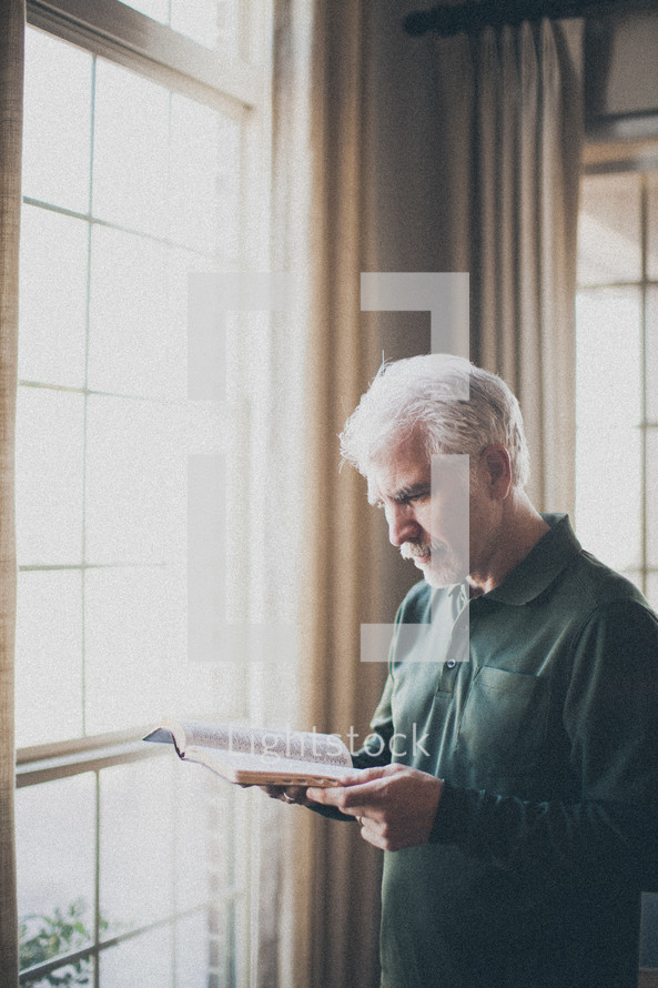 elderly man standing in front of a window Reading a Bible by sunlight