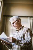 elderly man standing at a window reading a Bible
