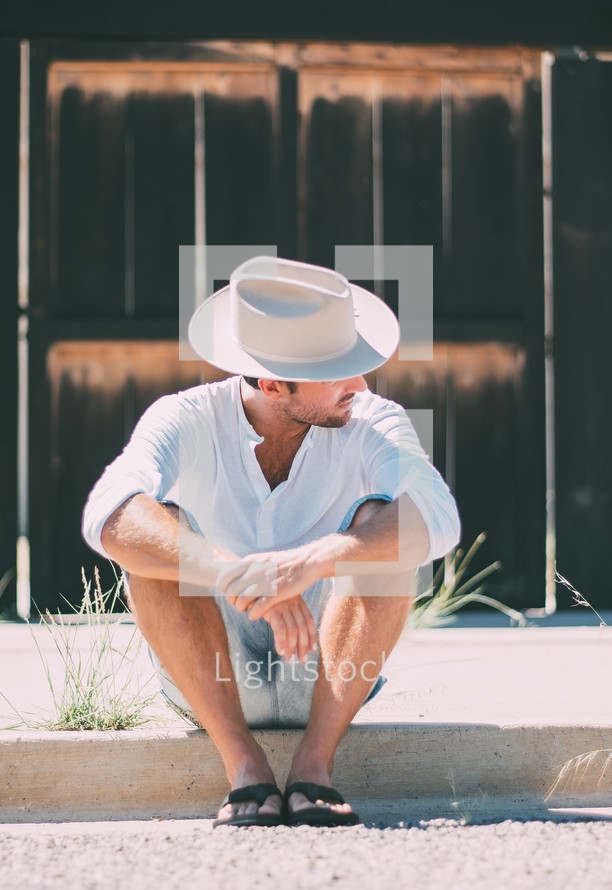 a man sitting on the curb in a cowboy hat and flip flops 