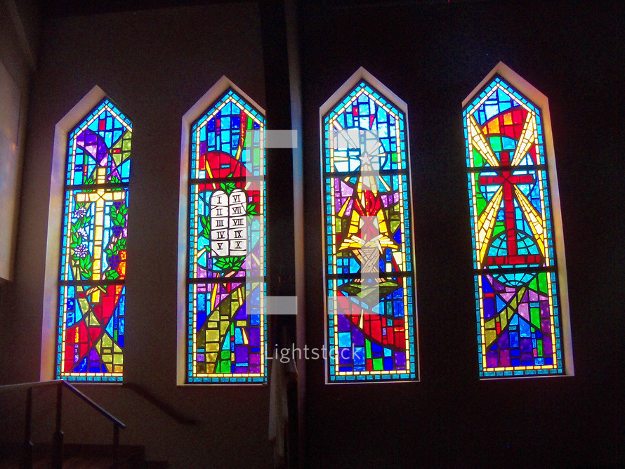 A set of four arched stained glass windows adorning a church with the cross, the ten commandments, The Manger nativity scene and resurrected power of Jesus.