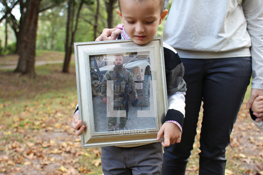 Boy standing in the woods with his mother, holding a framed photo of his soldier father.