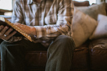 torso of an elderly man sitting on a couch reading a Bible