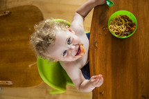 A little boy in a high chair eating spaghetti.