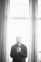 elderly man standing in front of a window praying by sunlight