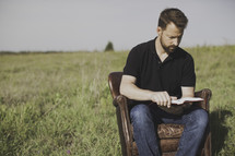 A man sitting in a leather chair reading a Bible outdoors 