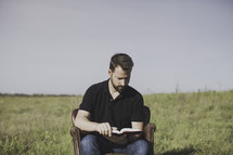 a man sitting in a leather chair outdoors reading a Bible 