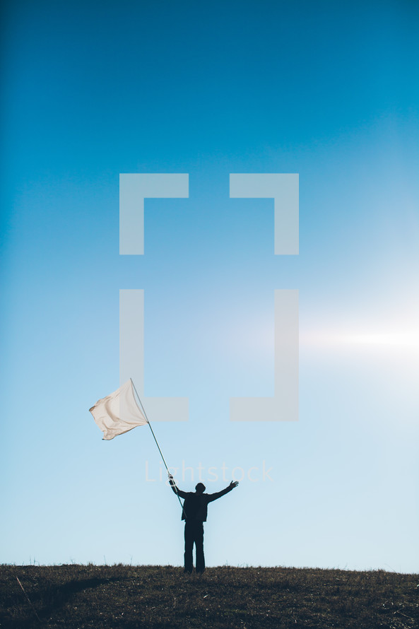 Silhouette of man waving white flag with arms raised on grassy hilltop.