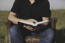 a man sitting in a leather chair outdoors reading a Bible 