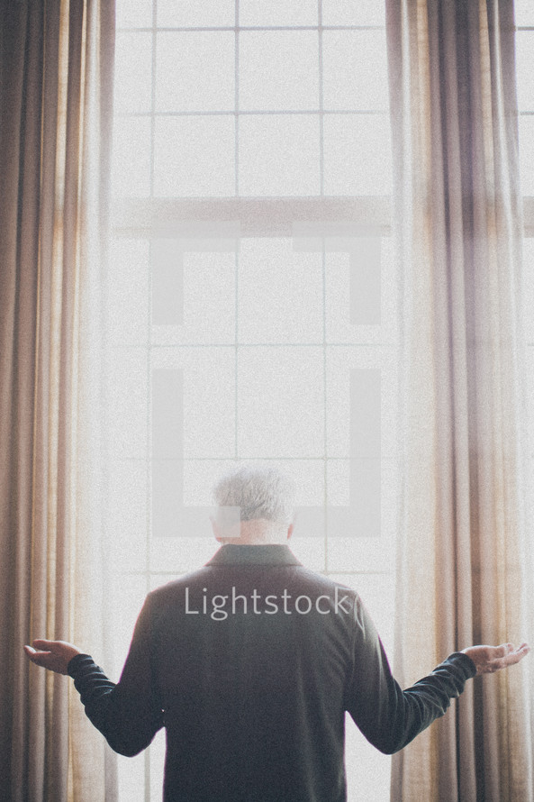 elderly man standing at a window with his arms outstretch in worship and praise to god