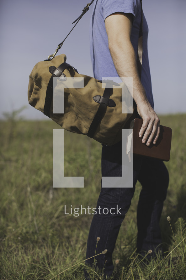 a man with a bag walking in a field of tall grass carrying a Bible 