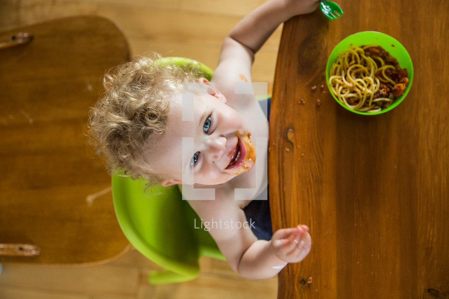 A little boy in a high chair eating spaghetti.