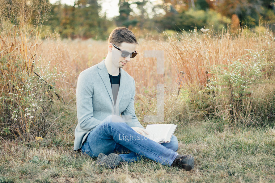 A man in a blazer sitting in the grass reading a Bible