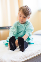 a boy child in a hospital gown and socks sitting  in a hospital bed 