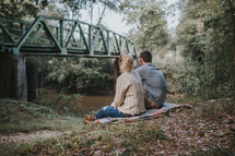 couple sitting on a shore looking at a bridge 