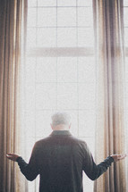 elderly man standing at a window with his arms outstretch in worship and praise to god
