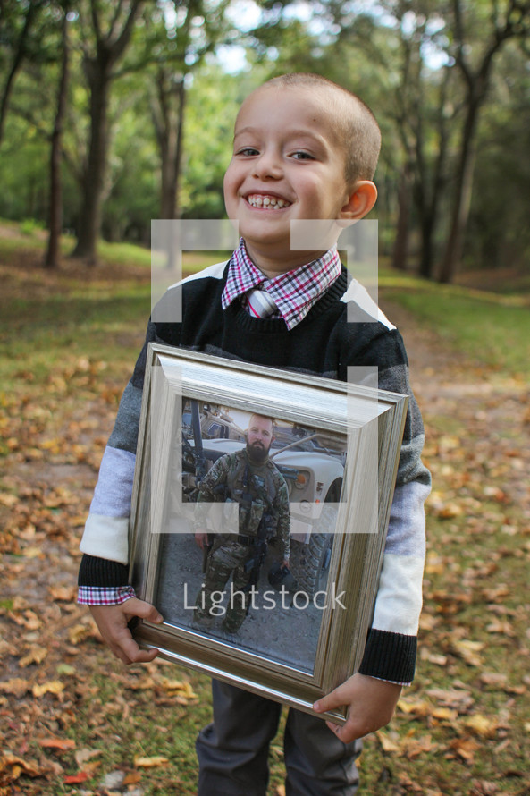 Young boy holding a picture of a soldier in camouflage
standing outside