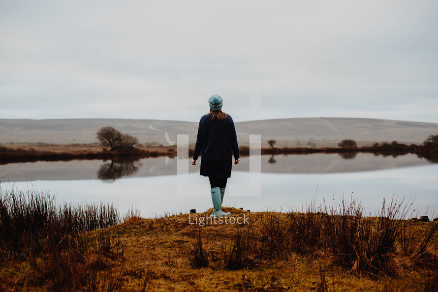 a woman in rain boots standing at the edge of a pond 